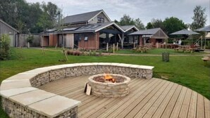 Ein Garten mit Feuerstelle aus Stein, Holzterrasse und einem Haus mit großer Garage und überdachter Außenbereich.