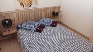 Een slaapkamer met een houten hoofdbord, een bed met een decoratief sprei, twee kussens en een plant aan de muur.