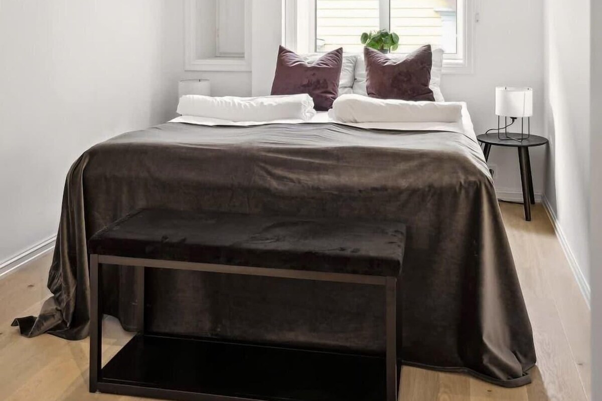 A bedroom with a bed, pillows, a bedside table, a lamp, and a window.