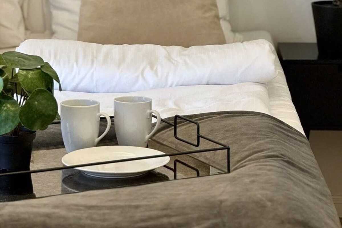 A neatly made bed with white bedding and a grey throw, a small table with a white plate and two mugs, and a potted plant to the left.