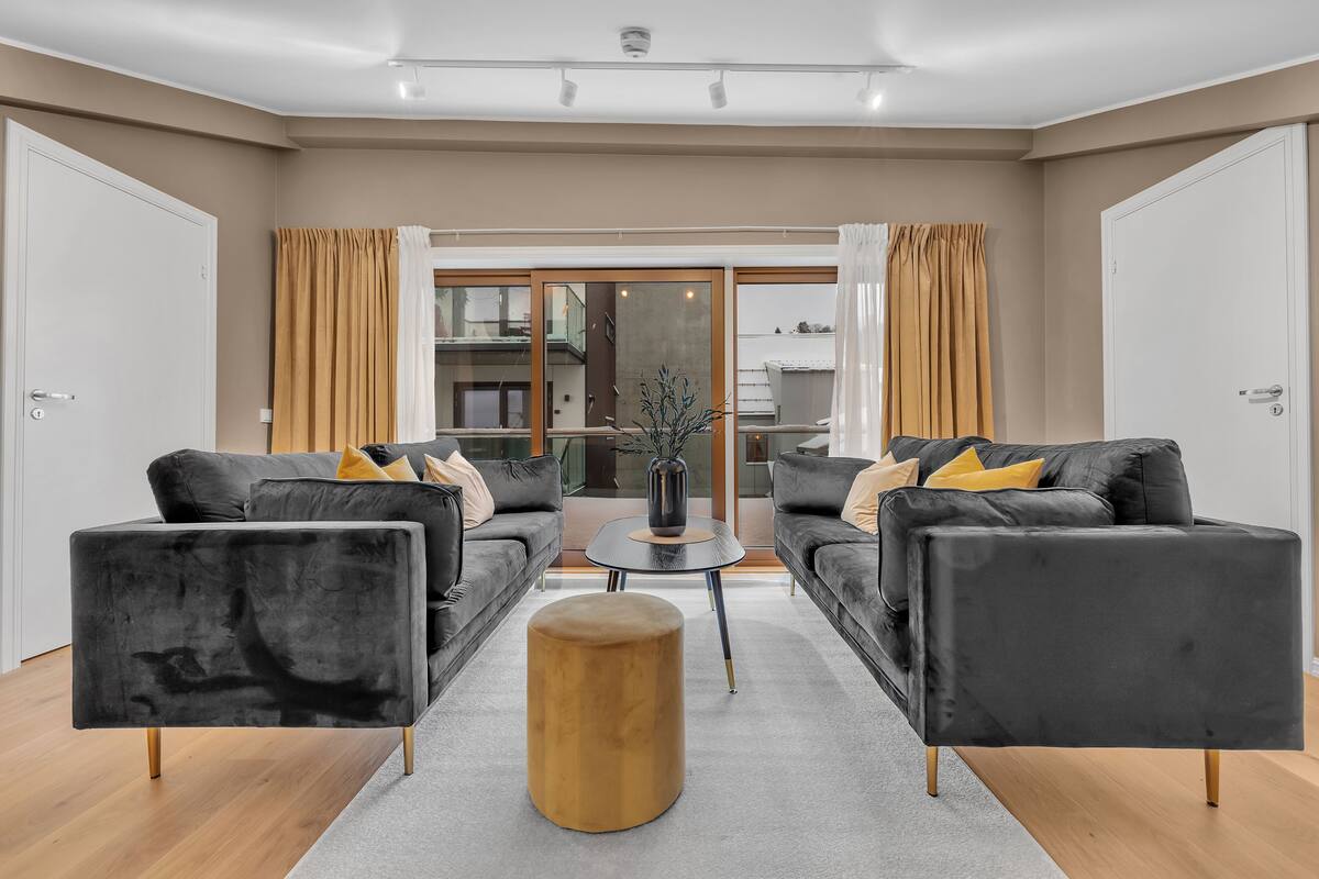 A modern living room with two black sofas, a round wooden ottoman, and a small table with a vase.