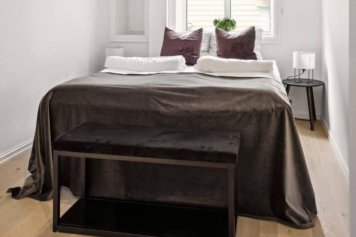 A bedroom with a bed, pillows, a bedside table, a lamp, and a window.