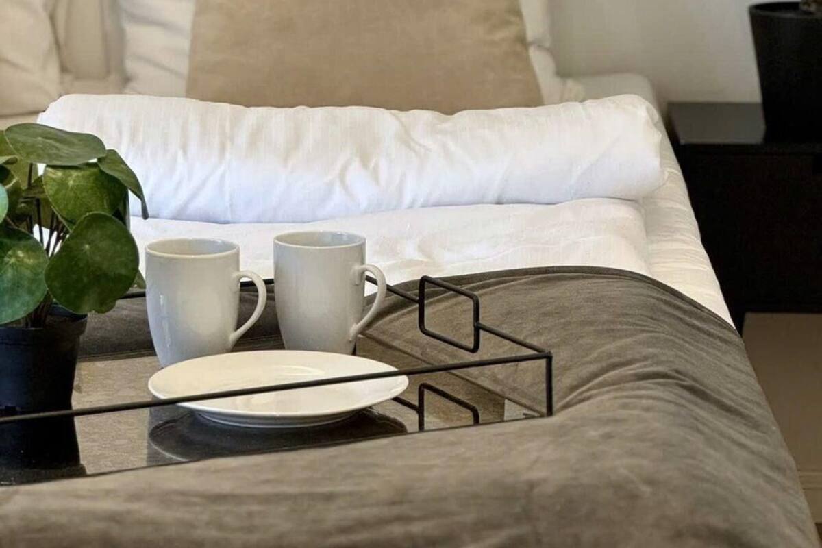 A neatly made bed with white bedding, a bedside table with a tray holding a plate, two cups, and a potted plant.