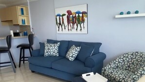 Un soggiorno con un divano blu, un poggiapiedi abbinato e una poltrona a motivi decorativi. Sul muro, un quadro raffigura persone con ombrelli.