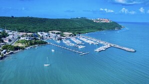 Vista aerea di una marina costiera con barche ormeggiate, una piccola barca a vela nell'acqua e un paesaggio collinare sullo sfondo.