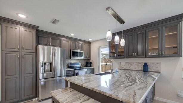 A modern kitchen with grey cabinets, stainless steel appliances, a marble countertop, and pendant lighting.