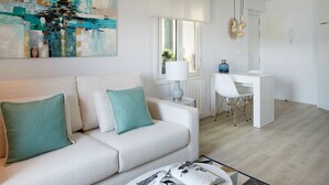 Ett modernt vardagsrum med en vit soffa, turkosa kuddar, ett soffbord med ett tidningsställ, ett skrivbord med en stol och en stor, abstrakt tavla.
