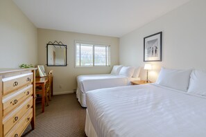 Une chambre d’hôtel avec deux lits, une commode en bois, un bureau, une chaise, une fenêtre avec des stores et un tableau encadré au mur.