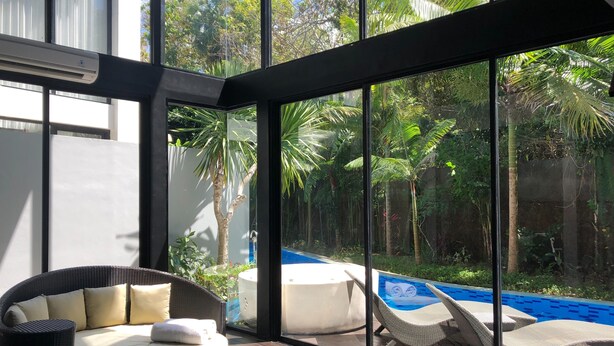 Una habitación moderna con un amplio ventanal, un sofá de mimbre, una piscina y abundante follaje exterior.