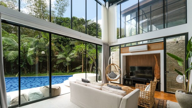 A modern living room with a large glass door leading to a pool, a sofa, a TV, and a wicker chair. - Inara Villa (Nusa Dua)
