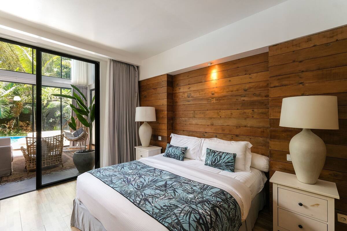 A bedroom with a large bed, wooden panel walls, two bedside lamps, a view of a swimming pool, and a potted plant.
