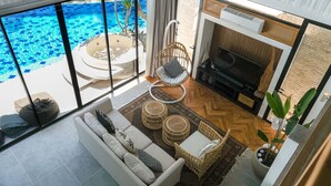 A modern living room with a large glass door leading to a pool, a sofa, a TV, and a fireplace. - Inara Villa (Nusa Dua)