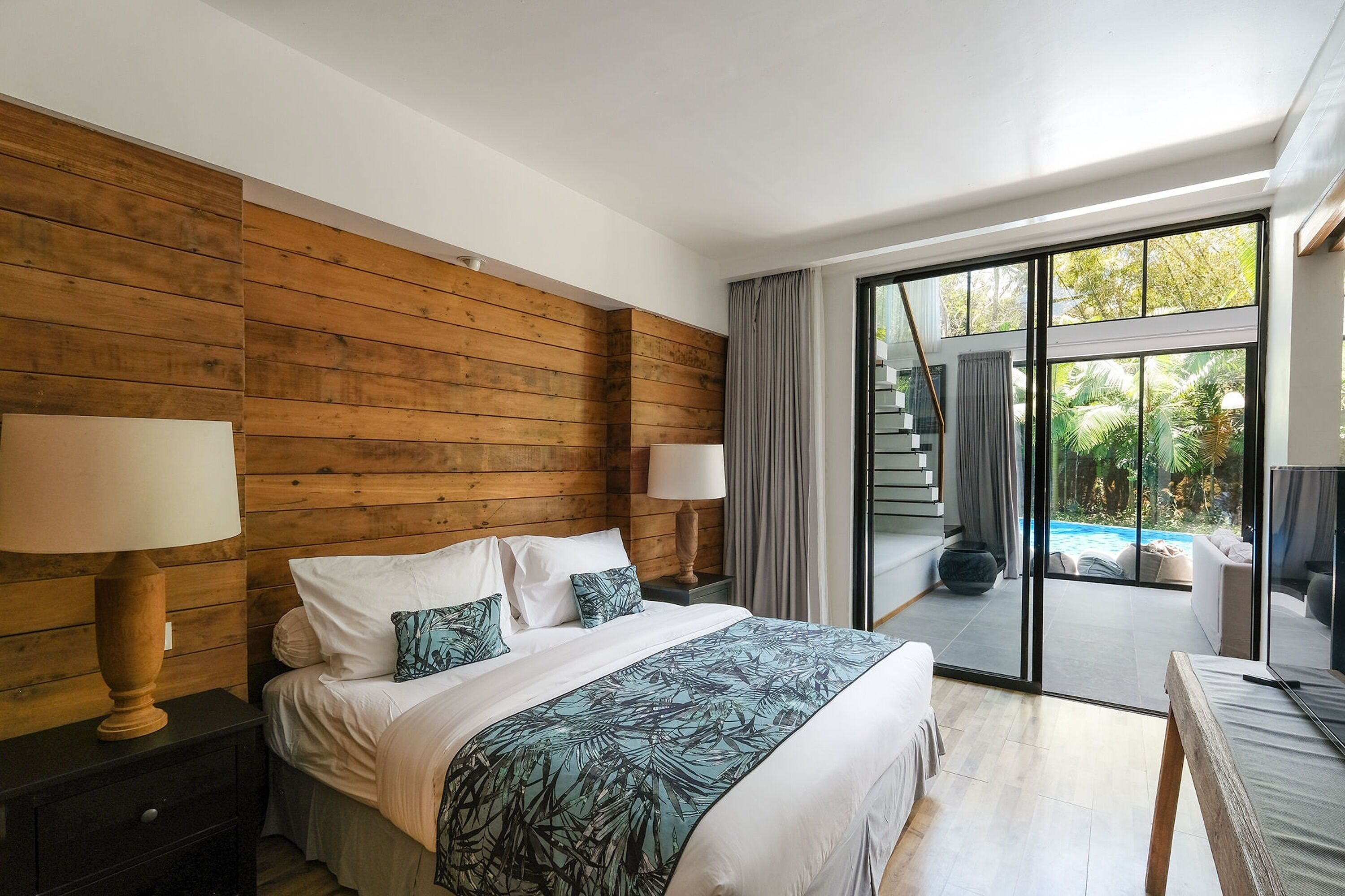 A bedroom with a wooden headboard, a large bed with white bedding and a tropical leaf-patterned throw, a nightstand with a lamp, and a view of a swimming pool through a glass door.