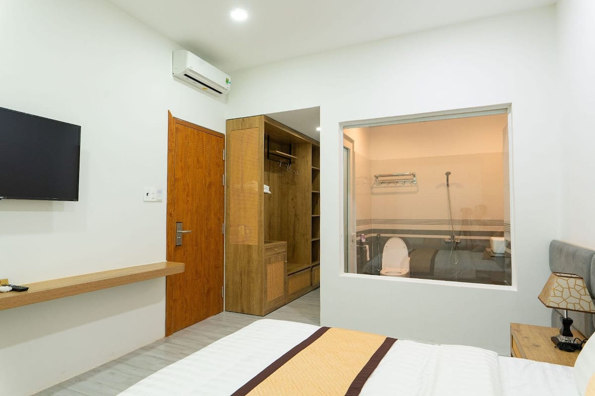 A bedroom with a bed, a TV mounted on the wall, a window with a view of a bathroom, and a wooden wardrobe.