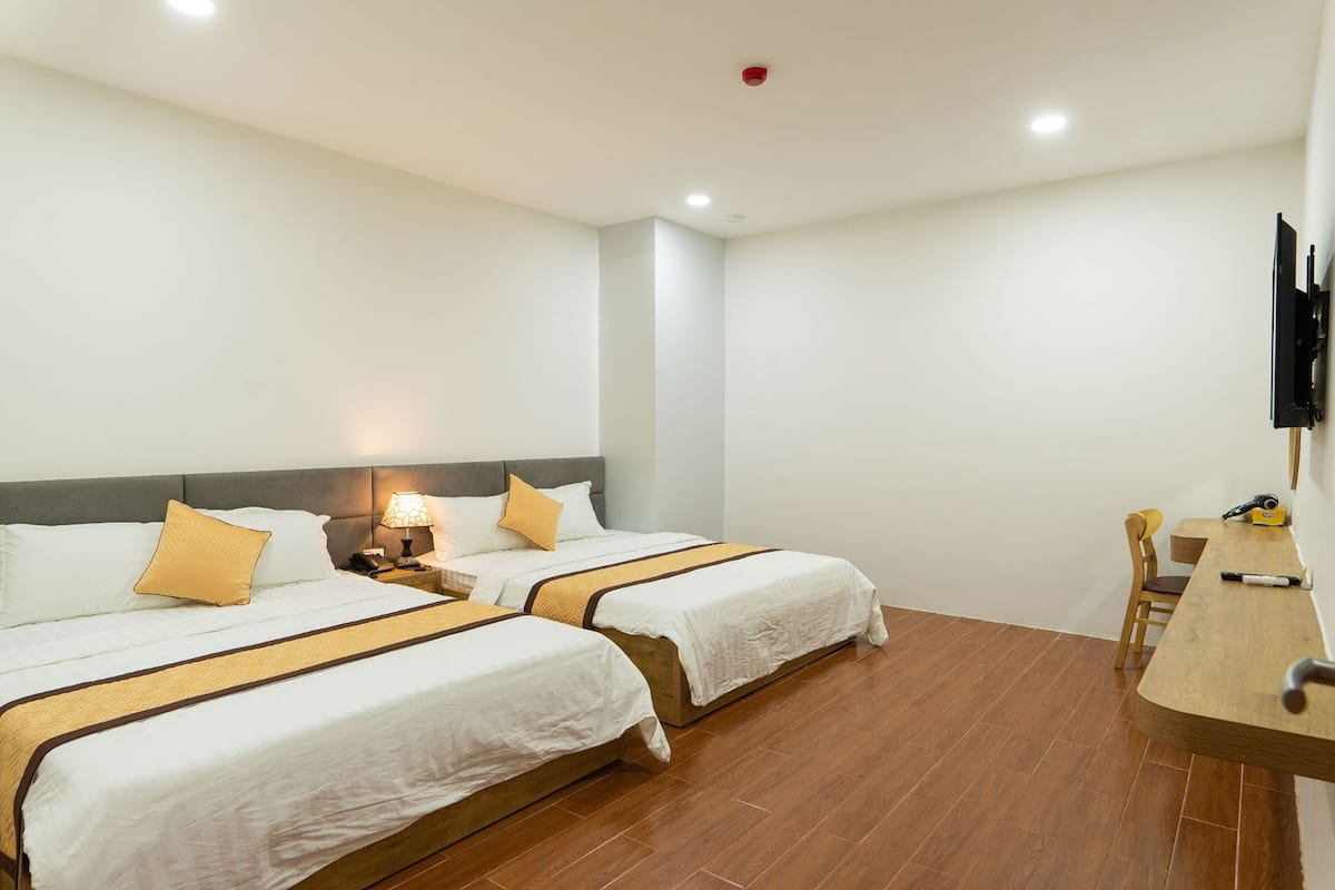 A hotel room with two beds, a desk, a chair, a TV, and a nightstand with a lamp.