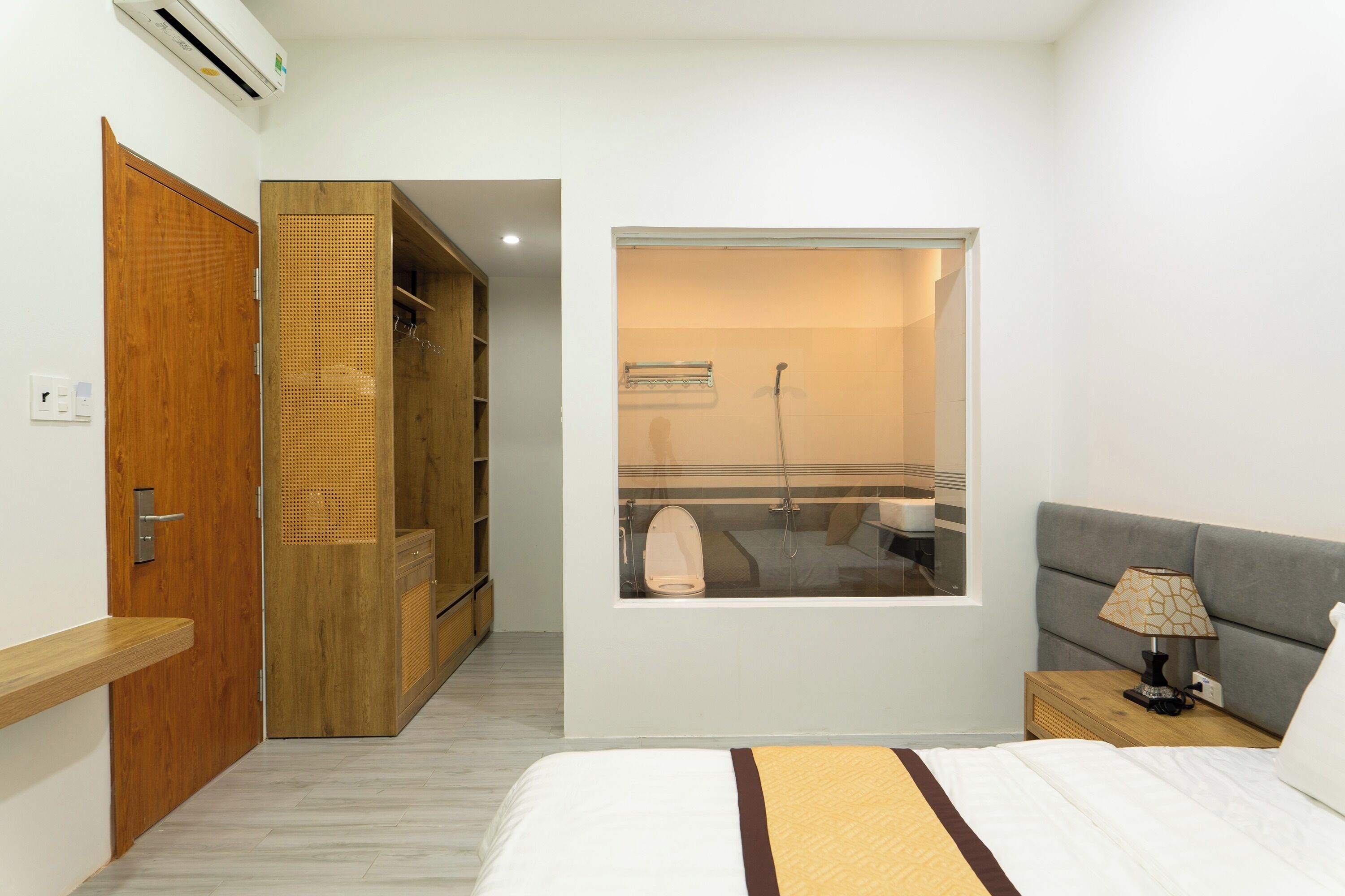 Un dormitorio con cama, mesita de noche, ventana, armario y baño visible a través de la ventana.