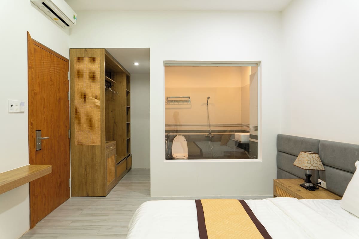 A bedroom with a bed, a nightstand, a window, a wardrobe, and a bathroom visible through the window.