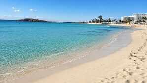 Ein Strand mit klarem türkisblauem Wasser, sandigem Ufer und einem Gebäude im Hintergrund.