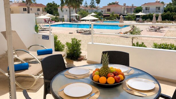 Ein runder Tisch mit Obstschale, Tellern und Besteck, unter einem großen Sonnenschirm auf einer Terrasse mit Pool und Liegestühlen im Hintergrund.