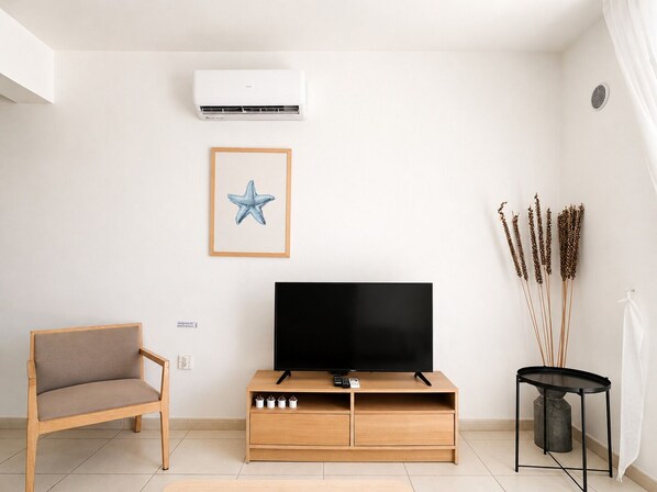 Un salon avec un téléviseur à écran plat posé sur un meuble en bois, une œuvre d’art représentant une étoile de mer encadrée, un fauteuil et une petite table agrémentée de quelques tiges décoratives.