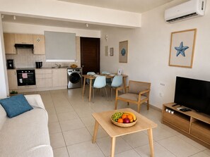 A compact living space with a kitchenette, dining area, and a seating area with a TV and a fruit bowl. - Narcissos 'Nissi Beach' Apartment C2 (Ayia Napa)