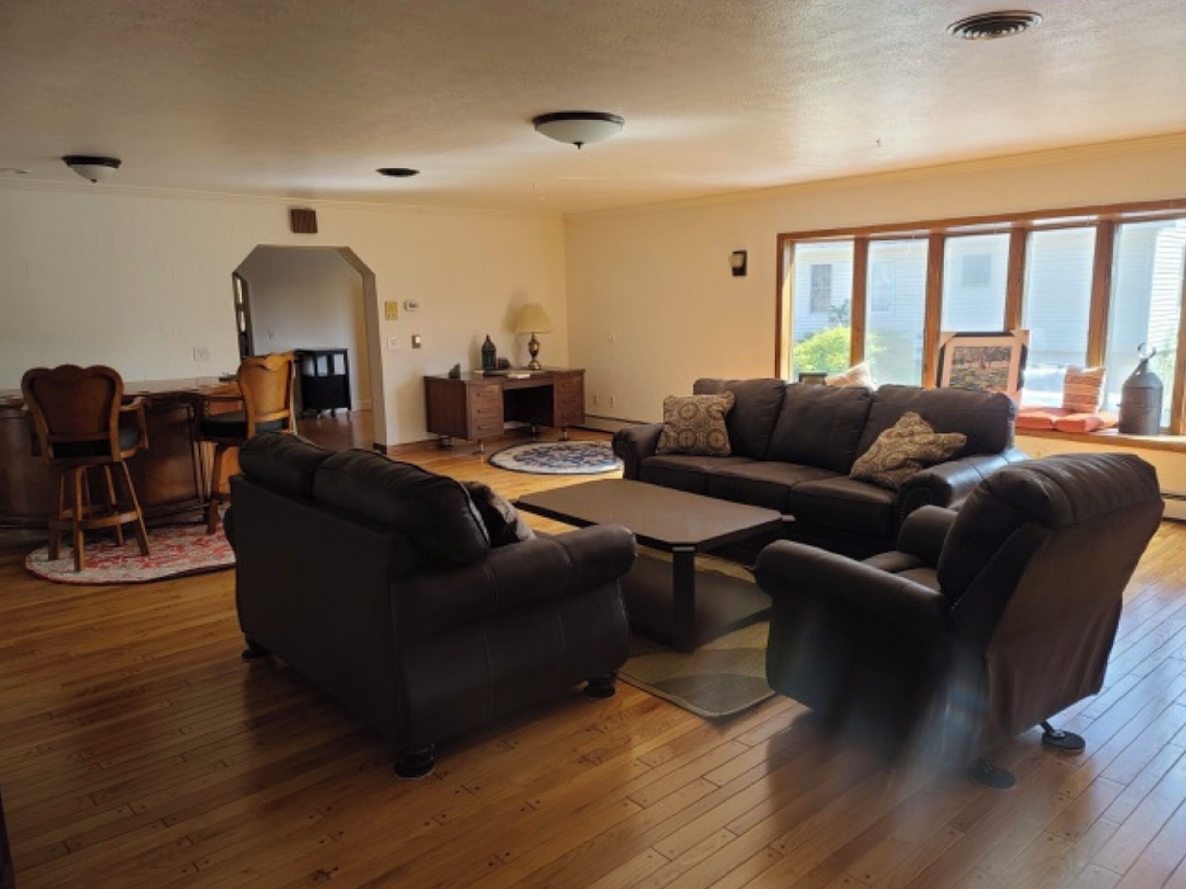 A spacious living room with a dark brown leather sofa, a coffee table, and a dining area with wooden chairs and a small desk.