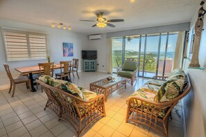Un amplio espacio de estar con mesa de comedor, mobiliario de mimbre, televisor y balcón con vistas panorámicas.