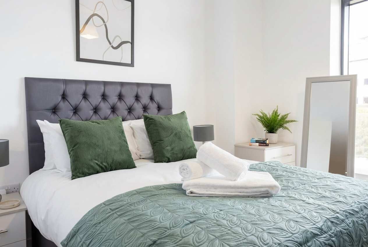 A neatly made bed with a green patterned bedspread and matching pillows, a bedside table with a lamp, and a small potted plant.