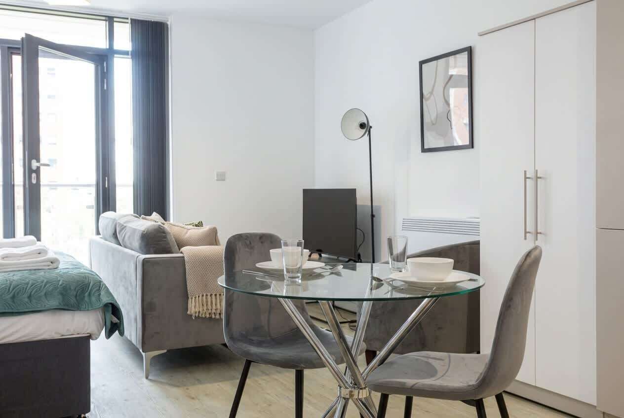 A modern hotel room with a glass dining table, grey upholstered chairs, a sofa, a flat-screen TV, and a wardrobe.