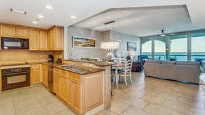 A modern kitchen with granite countertops, wooden cabinets, and a built-in microwave. There is a dining area with a chandelier and a view of the ocean through large windows.