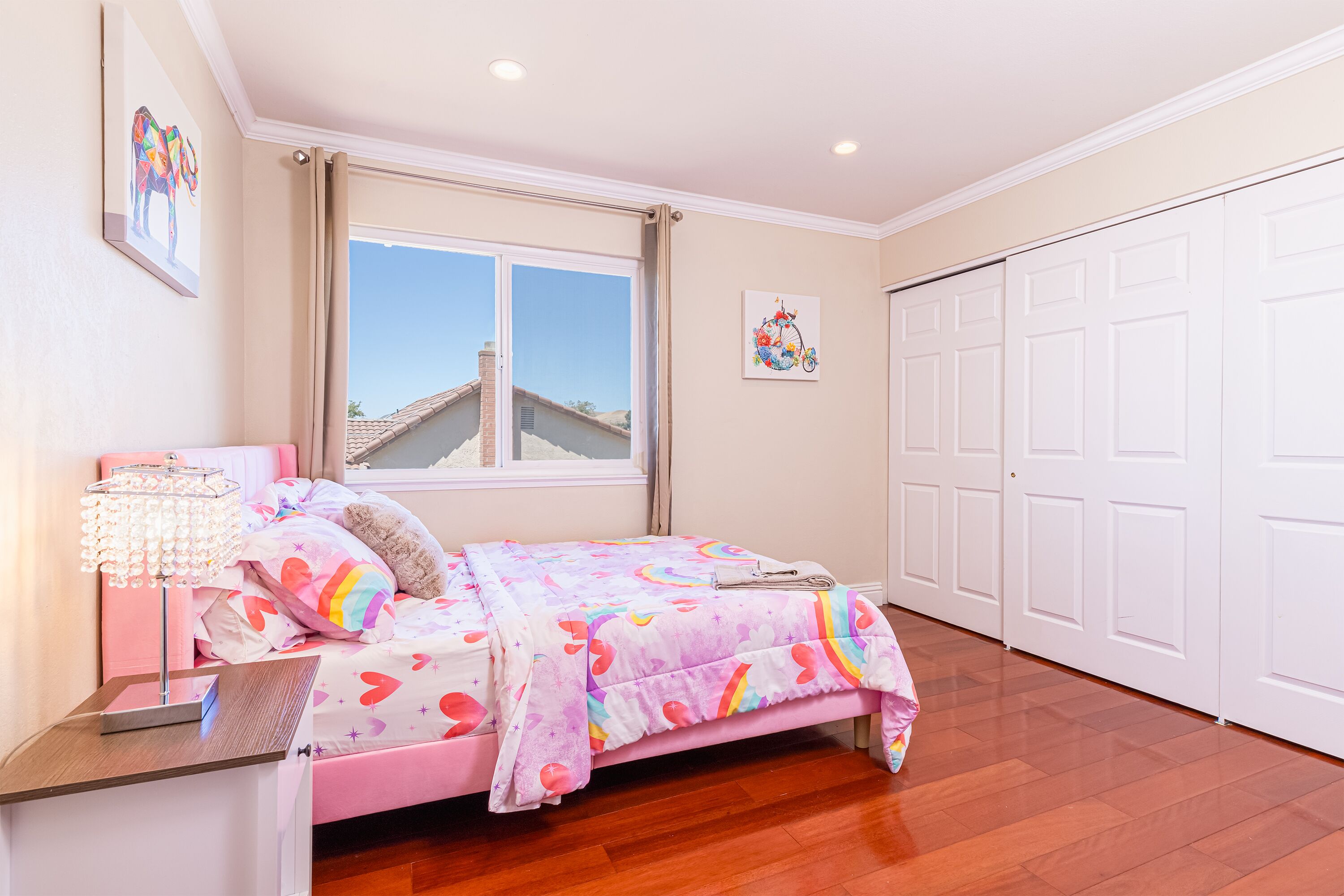 A bedroom with a pink bed, a nightstand, a lamp, and a window with a view of a house.