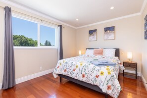 Un dormitorio con una cama grande, una mesita de noche, un ventanal con cortinas y dos cuadros colgados en la pared.