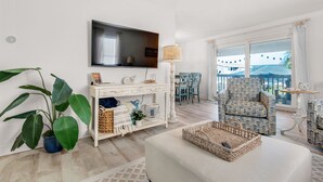 A modern living room with a large flat-screen TV, a white console table, a large green plant, a patterned armchair, and a view of the outside balcony.