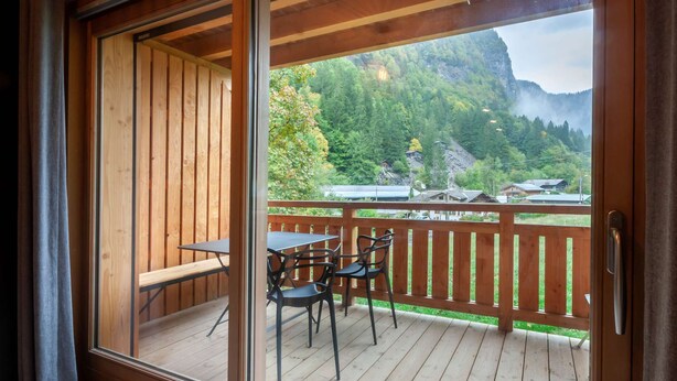 Una terraza de madera con una mesa y sillas, con vistas a un paisaje montañoso que incluye casas y un bosque.