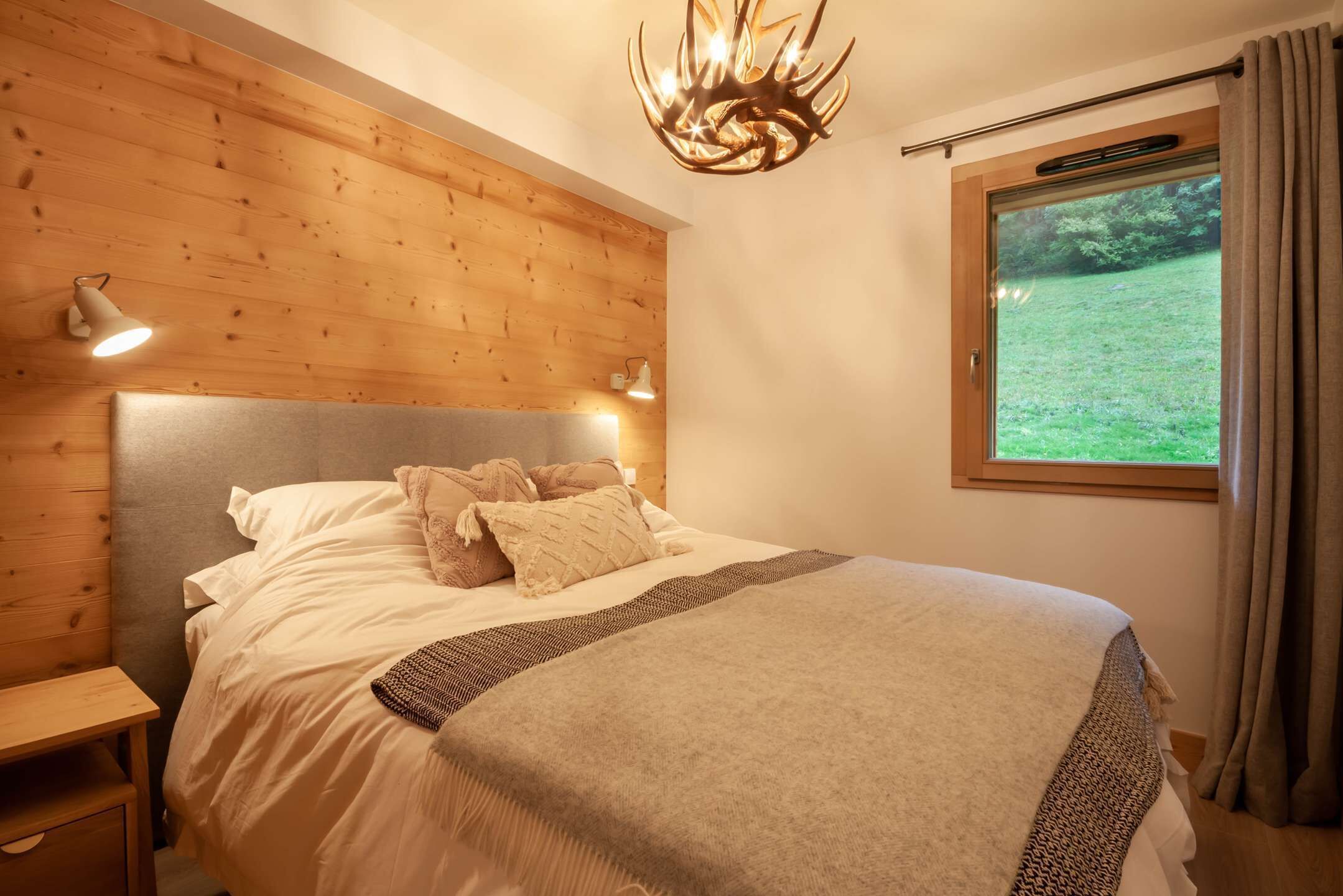 Un dormitorio con cabecera de madera, una cama grande con ropa blanca, dos lámparas de noche y una ventana con vistas a zonas verdes.
