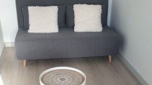 Um sofá cinzento com duas almofadas brancas e fofas, uma mesa redonda de madeira com tampo estampado e um tapete tecido sobre um chão de madeira clara.