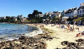 A beach with people swimming and sunbathing, rocky shoreline, and houses in the background.