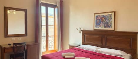 A hotel room with a bed, a desk, a chair, a mirror, a picture on the wall, and a window with curtains.