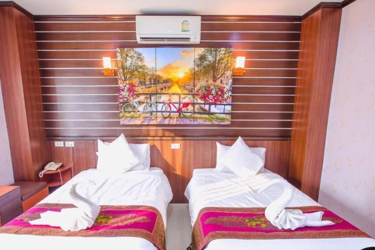 A hotel room with two beds, a painting of a sunset over a bridge, and an air conditioning unit above the beds.