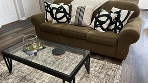 Ein Wohnzimmer mit einem Glastisch, einem braunen Sofa mit gemusterten Kissen und einem Teppich mit Struktur.