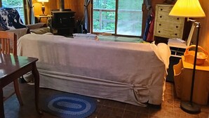 Una habitación acogedora con una cama cubierta con sábana blanca, suelo de madera, ventilador de techo, una mesa con lámpara y una ventana con cortinas.