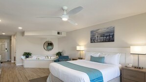 Une chambre d’hôtel moderne avec un grand lit, un ventilateur de plafond, une salle de bain avec un lavabo et un miroir, et un tableau accroché au mur.