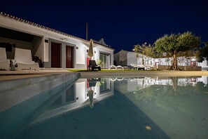 Una piscina con reflejo transparente, un edificio blanco con puerta roja y un árbol al fondo.