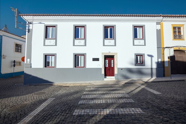 Un edificio blanco con puertas y ventanas rojas, una calle empedrada y un cielo azul despejado.