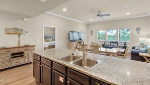 A spacious kitchen with granite countertops, a double sink, and wooden cabinets. There is a dining area with a table and chairs, and a living room with a sofa and a television.