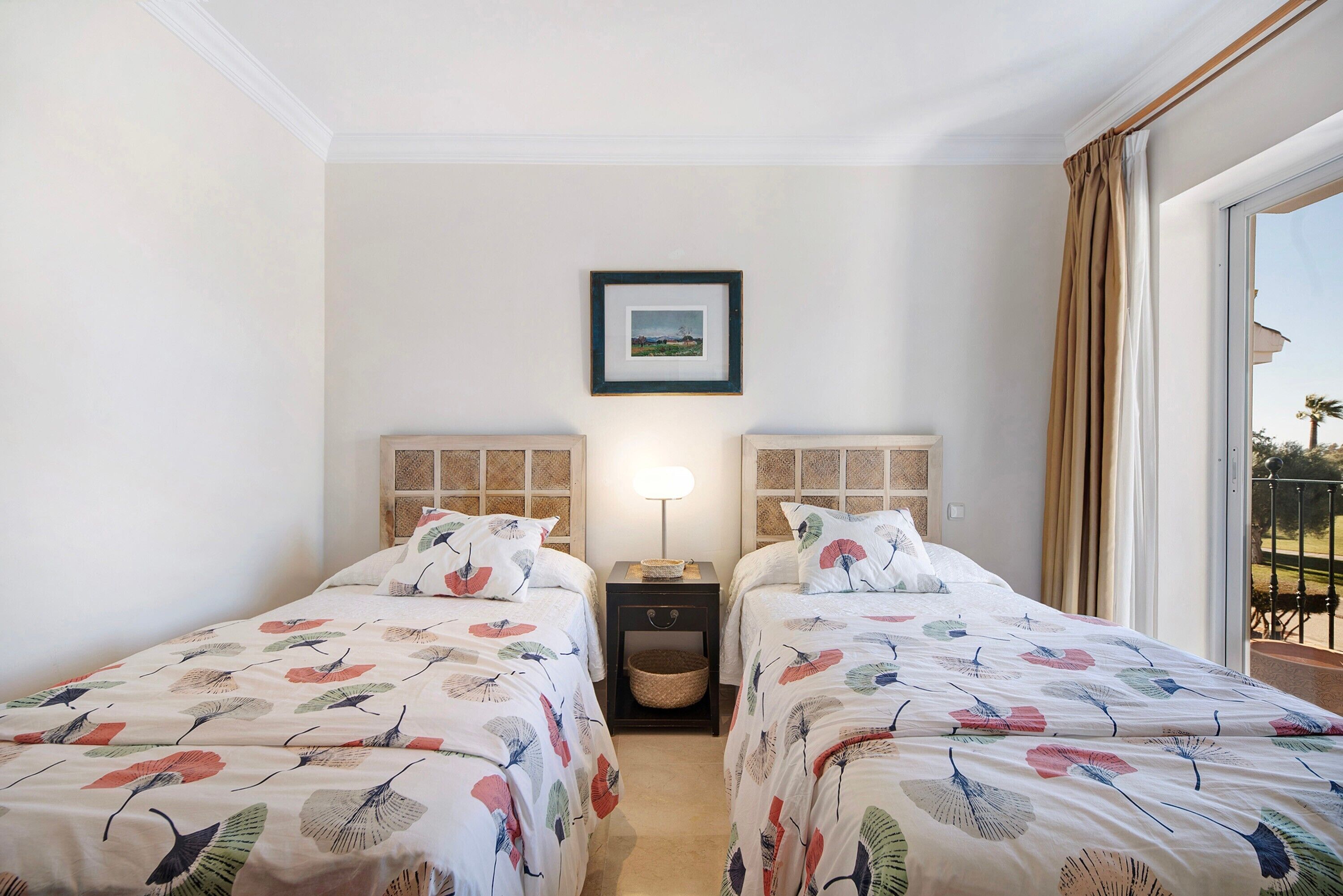 Hotellihuone, jossa on kaksi sänkyä, yöpöytä lampun kanssa, seinällä kehystetty taulu ja näkymä ulos.