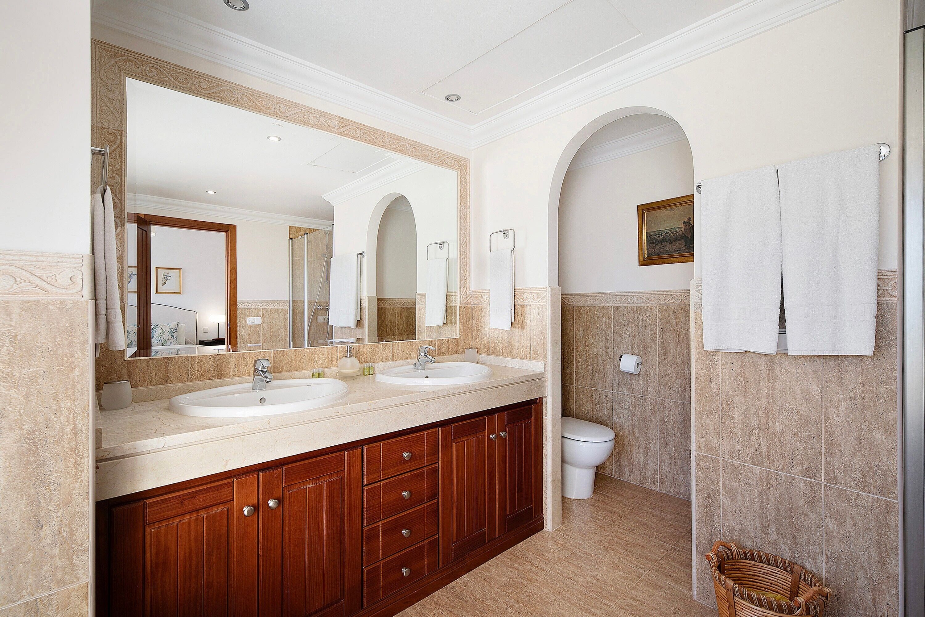 A bathroom with two sinks, a large mirror, wooden cabinets, a white toilet, and a wicker basket.
