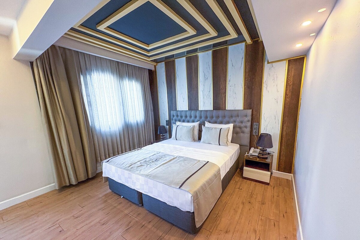 A modern hotel room with a large bed, wooden flooring, a striped wall design, and a ceiling with geometric patterns.