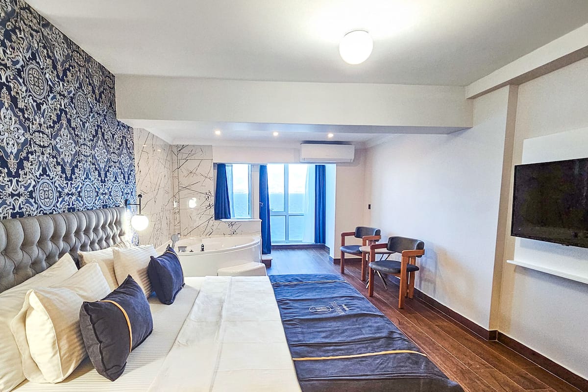 Une chambre d’hôtel moderne dotée d’un grand lit, d’un coin salon avec une table et des chaises, d’une grande fenêtre offrant une vue agréable et d’un téléviseur à écran plat fixé au mur.