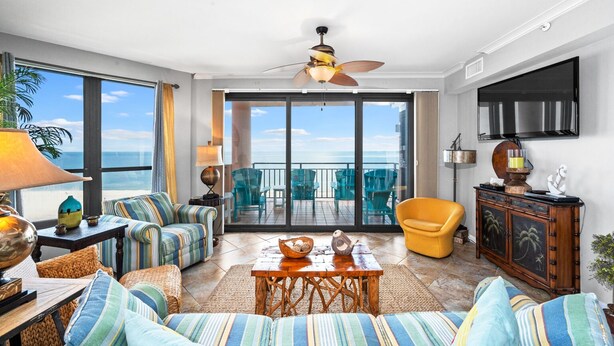 Een ruime woonkamer met een balkon en uitzicht op de oceaan, met een plafondventilator, een televisie en een houten kast.